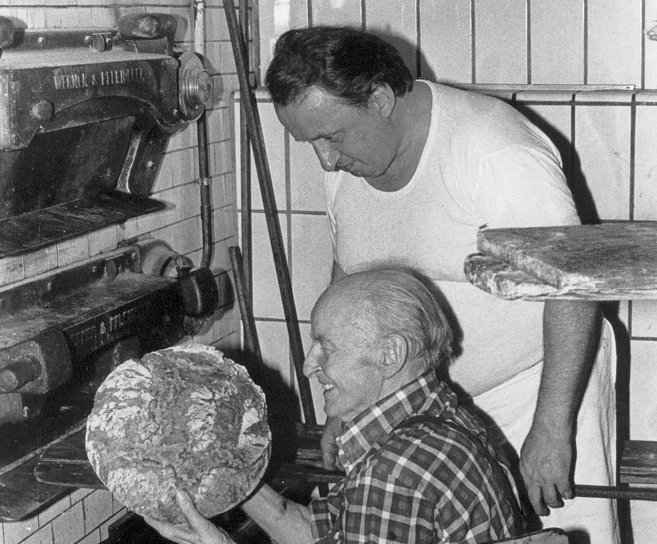 Historische Schwarzweiß-Aufnahme von Wilhelm Pappert, Gründer der Bäckerei Pappert, der ein Brotlaib in seinen Händen hält. Im Hintergrund ist auch Sohn Josef Pappert zu sehen.