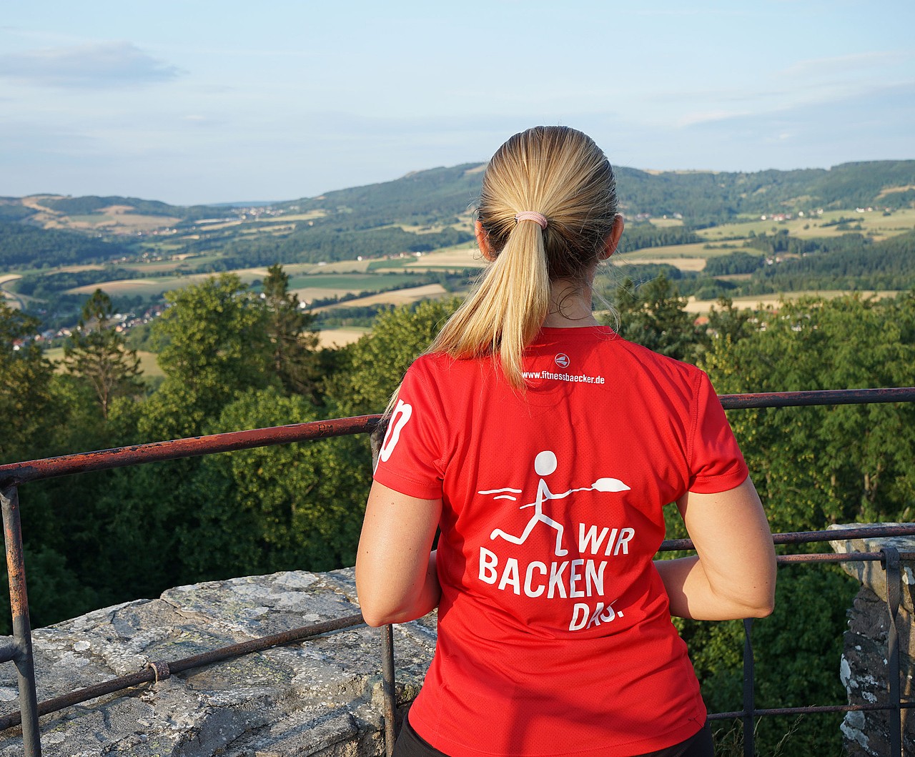 Fitnessbäcker-Teilnehmerin mit rotem T-Shirt mit der Aufschrift „Wir backen das“, blickt von einer Aussichtsplattform auf eine grüne Landschaft.