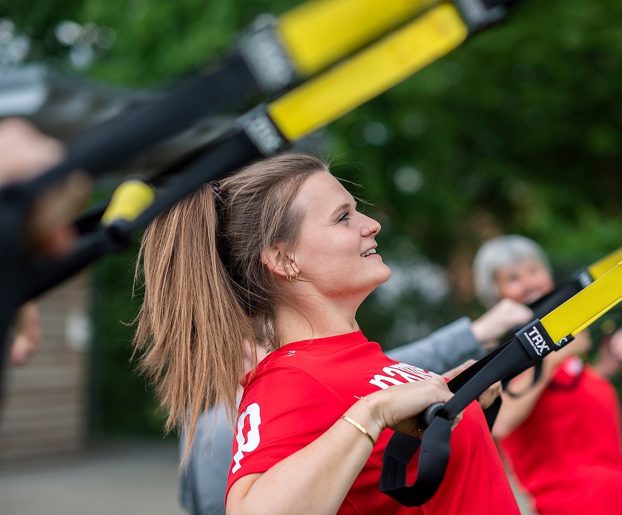 Fitnessbäcker Kurs-Teilnehmerin in rotem Pappert-T-Shirt trainiert im Freien mit TRX-Bändern; im Hintergrund sind weitere Teilnehmende des Fitnesskurses zu sehen.