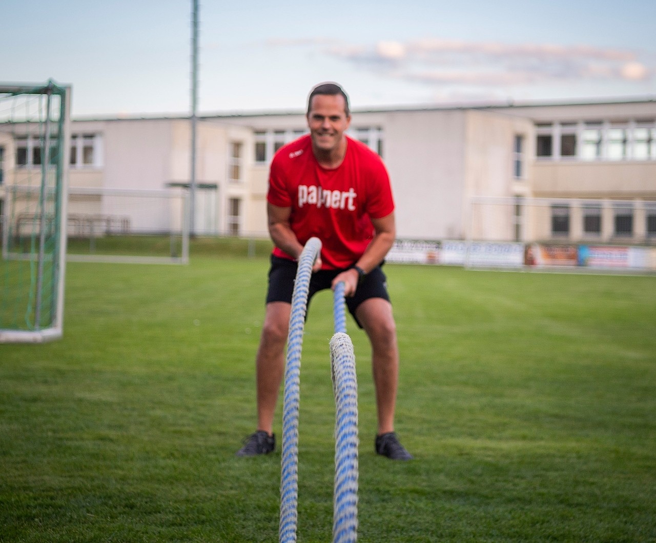 Ein Fitnessbäcker Kurs-Teilnehmer trainiert im roten Pappert-T-Shirt im Freien auf einer Wiese mit Battle Ropes und lächelt dabei in die Kamera.