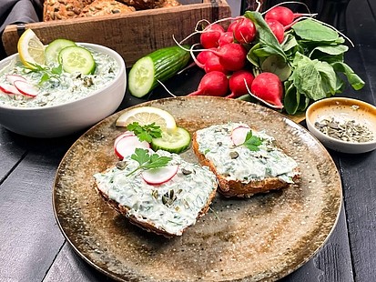Frischer Kräuterfrischkäse mit Gurke, Radieschen und Petersilie, serviert auf dem KürbisFIT-Brötchen sowie platziert auf einem edlen Teller.