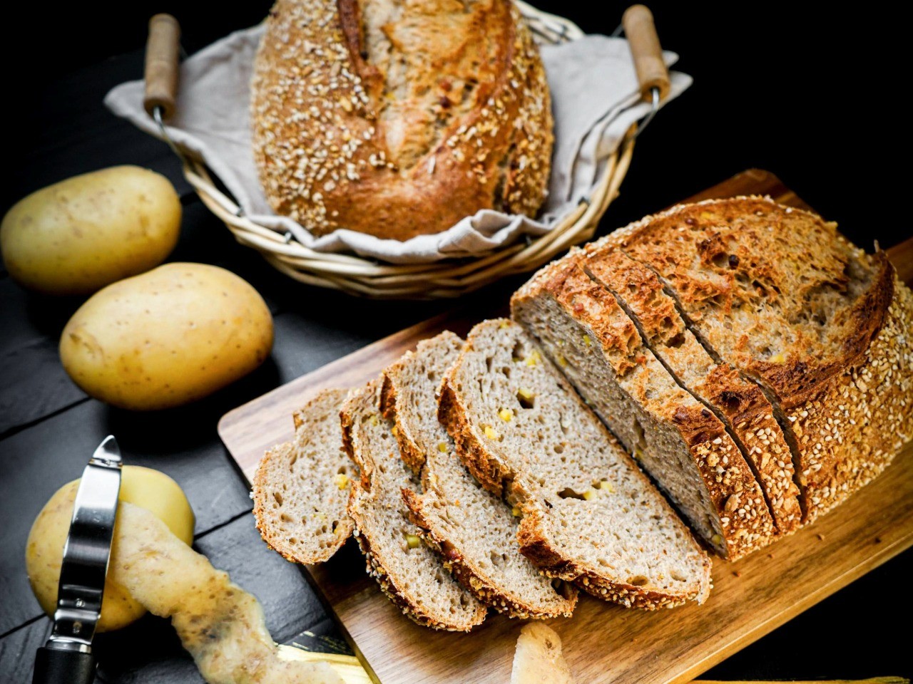 Das Kartoffelbrot der Bäckerei Pappert mit Kartoffelstückchen. Das Brot liegt aufgeschnitten und am Stück auf dunklem Holzhintergrund. Daneben liegen frische Kartoffeln und ein Kartoffelschäler.