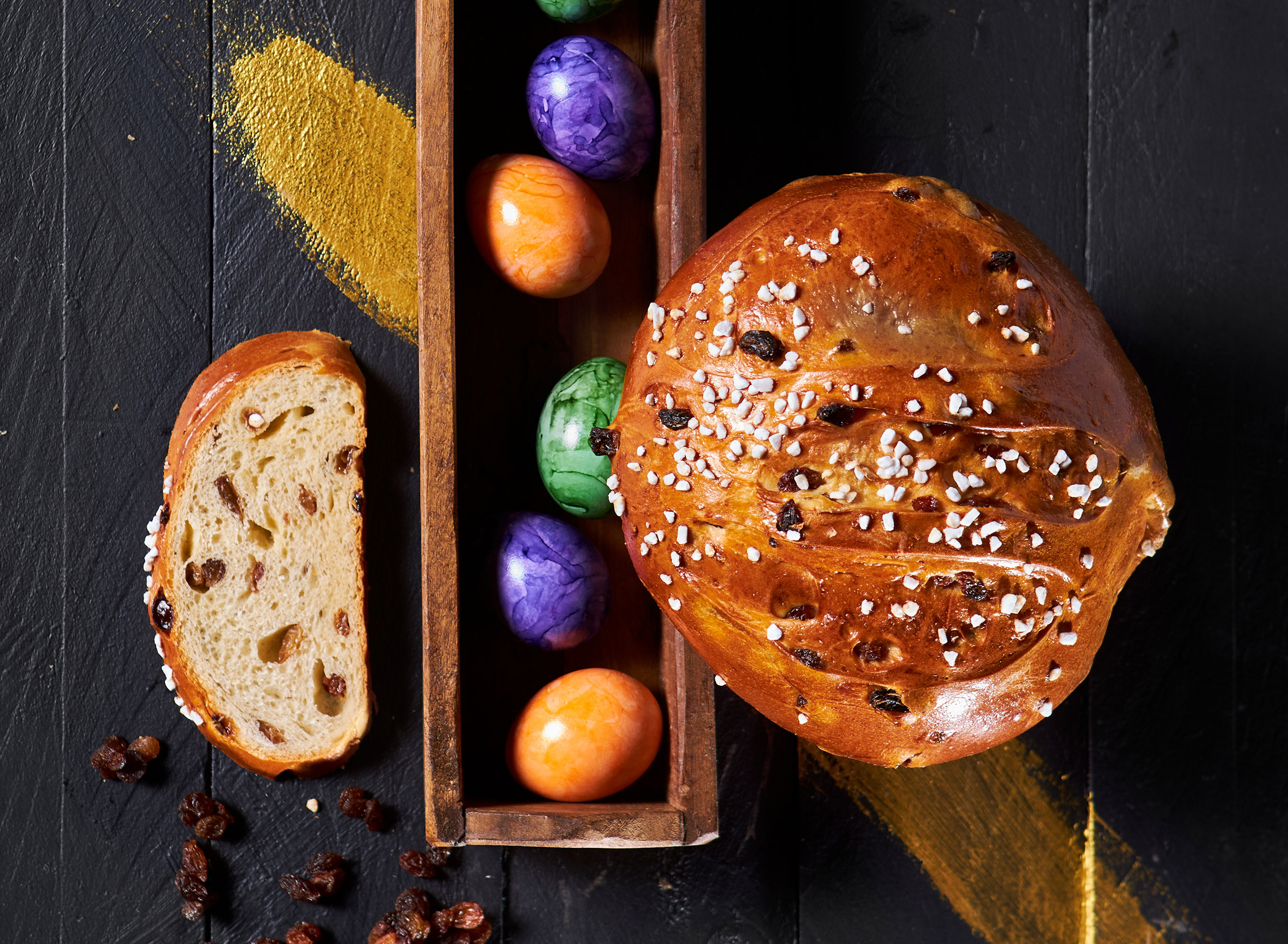 Das Osterbrot der Bäckerei Pappert mit mehreren bunten Ostereiern auf einem schwarzem Holzhintergrund platziert. 