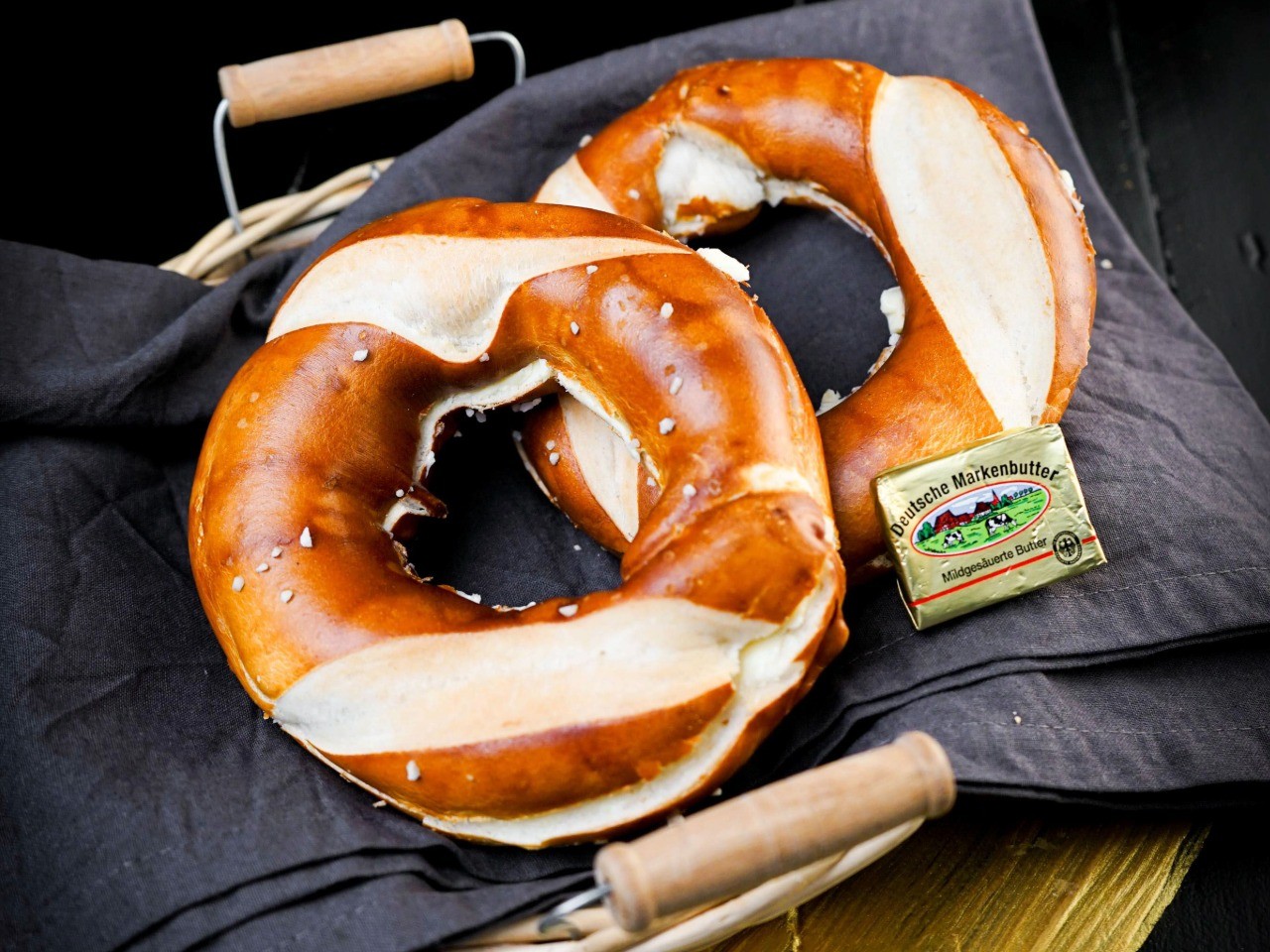 Der Butter-Laugenring der Bäckerei Pappert auf schwarzem Holzhintergrund. Dekoriert mit Tuch, Korb und einem Stück Butter.