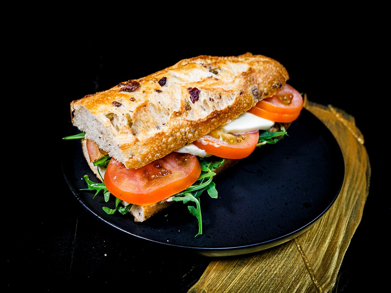 Der Oliven Franz Tomate-Mozzarella der Bäckerei Pappert auf schwarzem Teller, platziert auf einem dunklem Holzhintergrund.